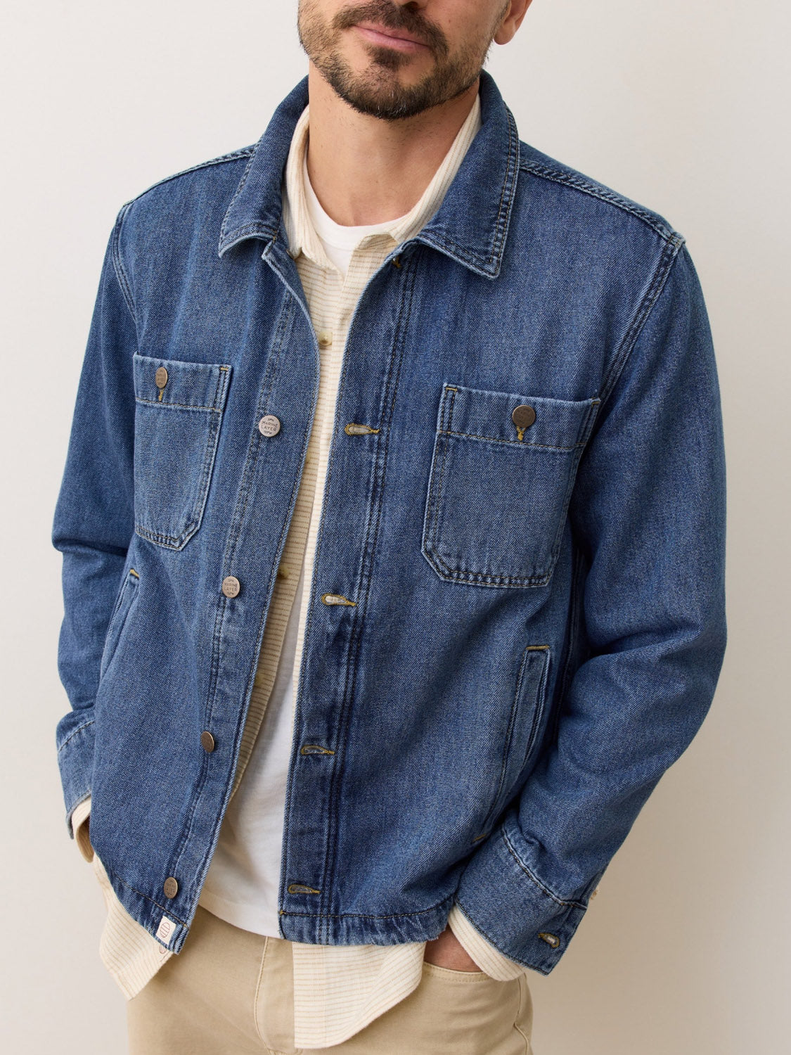 A man stands with his hands in his pockets against a plain background, wearing Marine Layer's Patrick Denim Overshirt in blue 100% cotton over a cream textured shirt and beige pants.