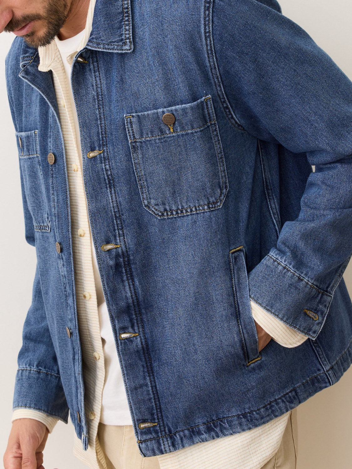 A man wears the Marine Layer Patrick Denim Overshirt over a cream button-up shirt and beige pants, with his left hand in his pocket; only his lower face and torso are visible.