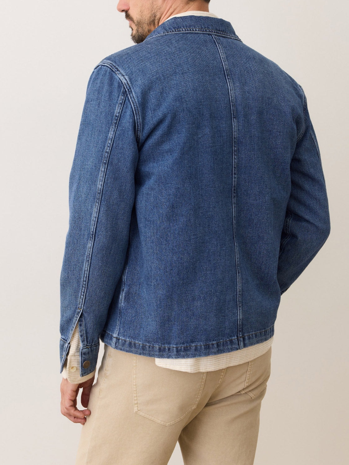 A man is shown from the back, wearing the Marine Layer Patrick Denim Overshirt in blue with beige pants, standing against a plain light background.