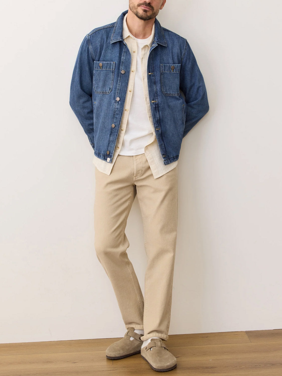 A man stands against a plain wall wearing the Marine Layer Patrick Denim Overshirt (100% cotton) over a white shirt, beige pants, and tan suede shoes. His hands are in his jacket pockets, standing on a wooden floor.