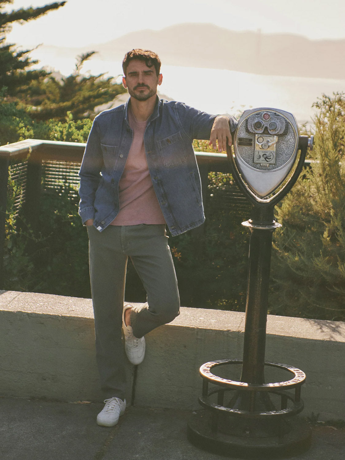 A man in the Marine Layer Patrick Denim Overshirt, pink shirt, gray pants, and white sneakers leans against a coin-operated viewer outdoors with scenic trees, water, and hills in the background.