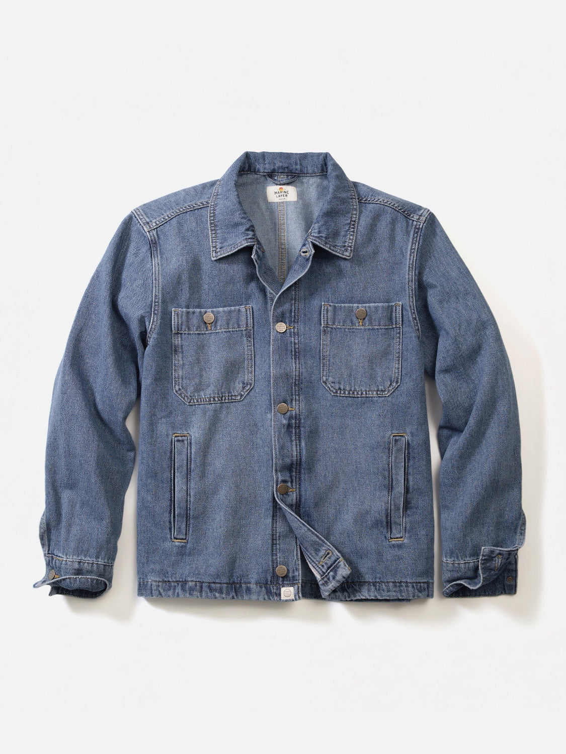 The Patrick Denim Overshirt by Marine Layer is a light blue, 100% cotton overshirt with long sleeves, a button-up front, two chest pockets with button flaps, and two side pockets, shown laid flat on a white background.