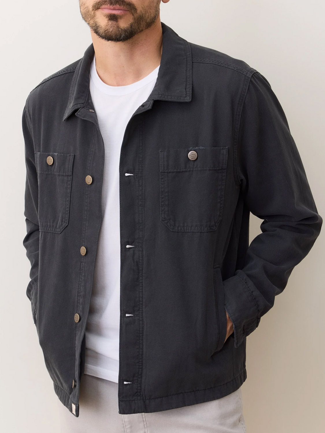 A man wears the Marine Layer Patrick Canvas Overshirt over a white t-shirt and light pants, standing with hands in his pockets against a plain light background.