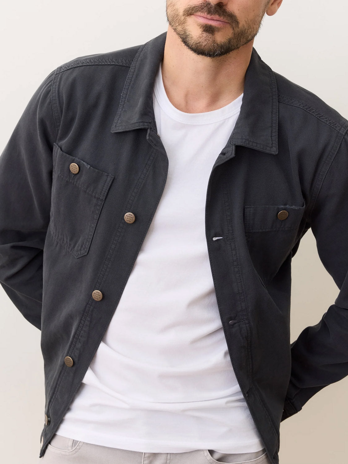 A man models the Marine Layer Patrick Canvas Overshirt in dark fabric over a white t-shirt, standing against a light background with his hands behind his back; only his torso and lower face are visible.