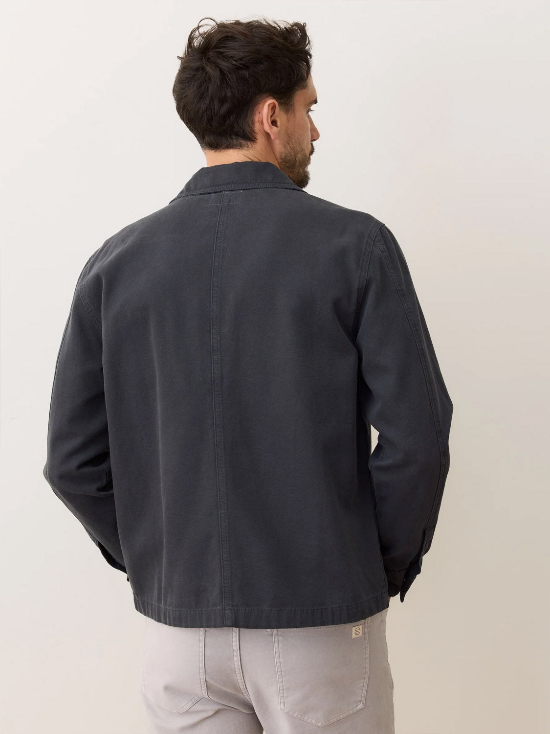 A man with short, dark hair stands with his back to the camera, wearing the Marine Layer Patrick Canvas Overshirt in dark gray and light gray pants, set against a plain, light background.
