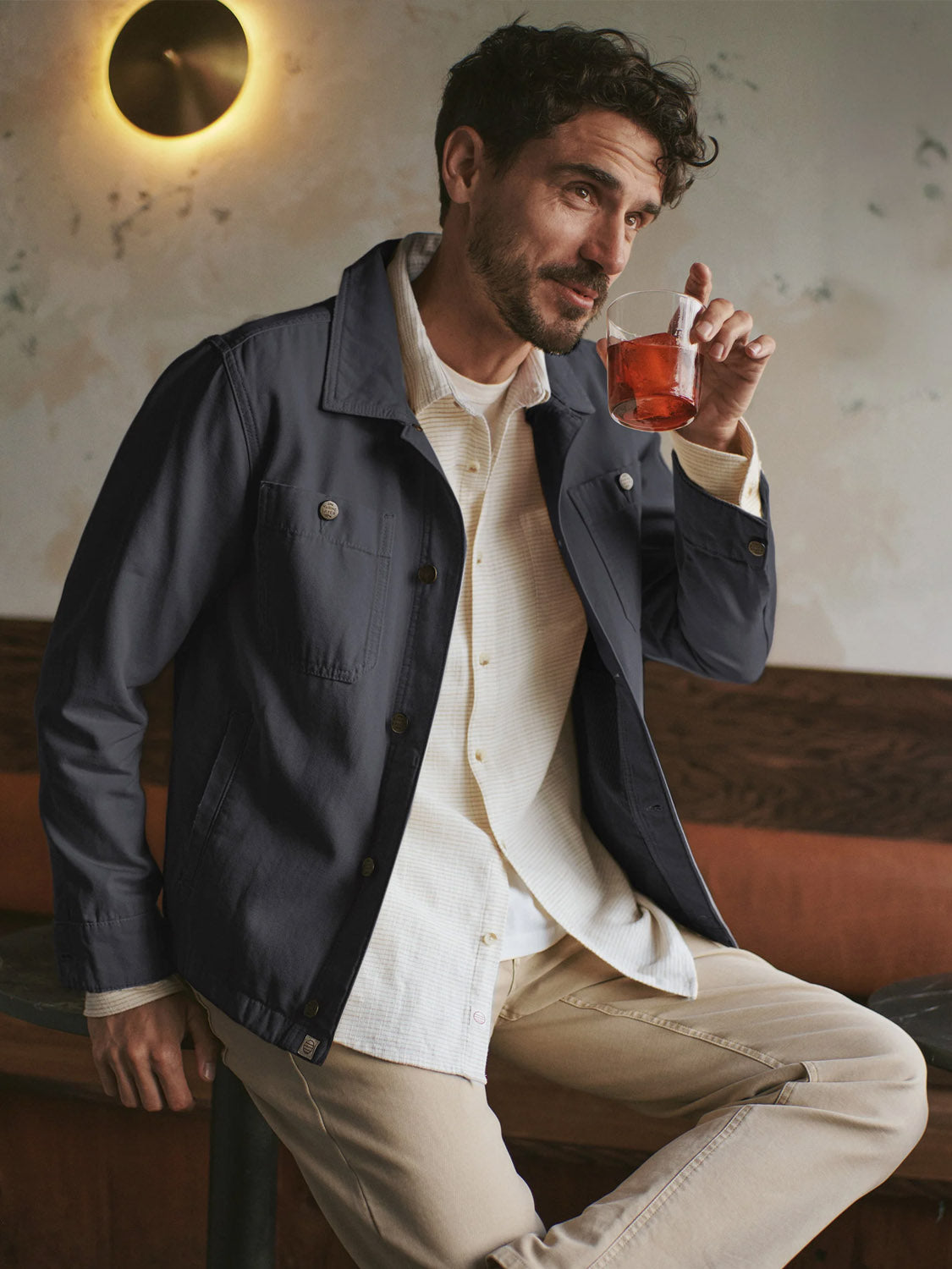A man with dark curly hair and a beard sits on a bench, sipping a red drink, wearing Marine Layer’s Patrick Canvas Overshirt over a light shirt and khaki pants, with a round wall light glowing in the background.