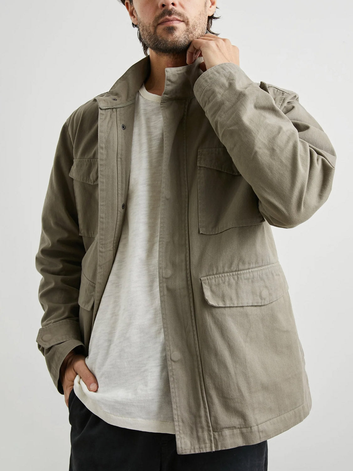 A man faces the camera against a plain background, wearing the Rails Paulsen Jacket in light olive green over a white t-shirt and black pants, with one hand touching his collar.
