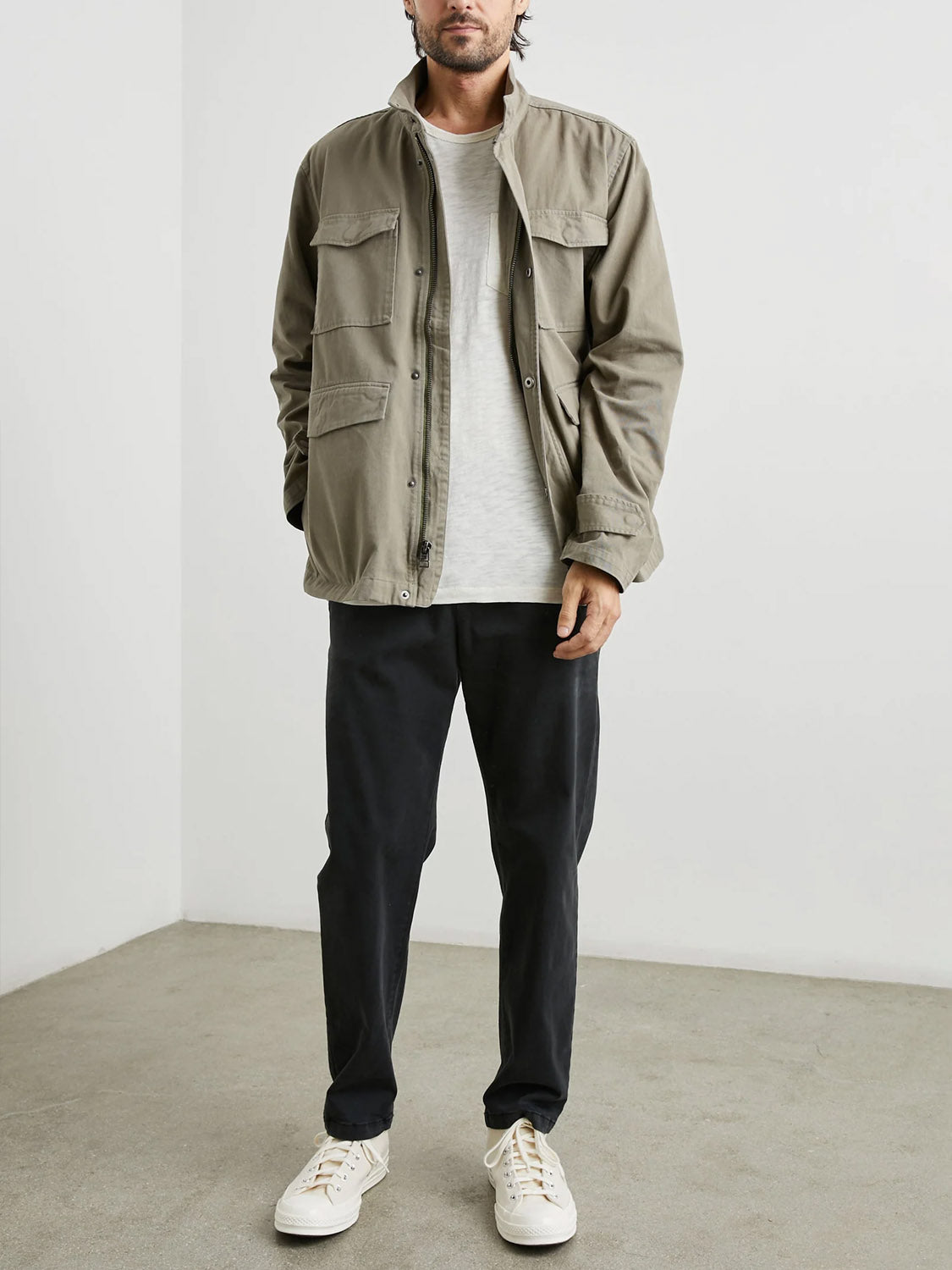 A man stands indoors facing forward, wearing the Rails Paulsen Jacket in light olive over a white t-shirt, black pants, and white sneakers. He has a neutral expression and the background is plain and minimal.