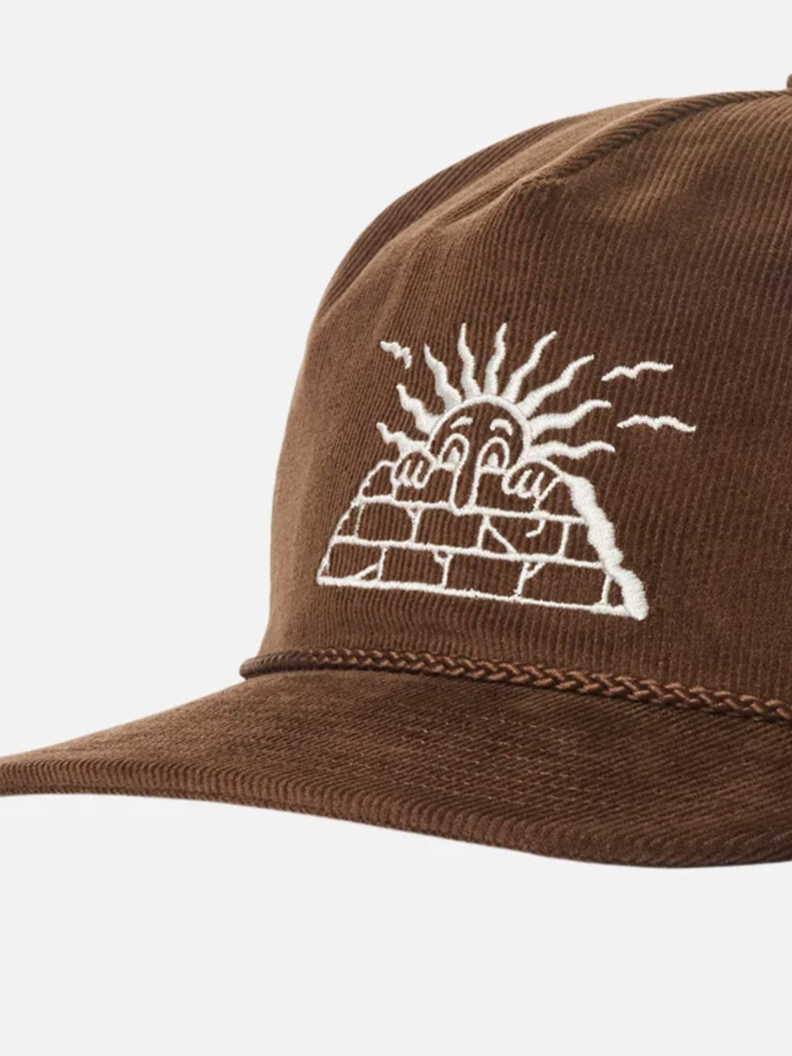 The Katin Peaks Hat is a brown cotton corduroy 5-panel cap with white stitching, featuring an embroidered face over a brick wall with radiating lines, and finished with an adjustable snapback for comfort.