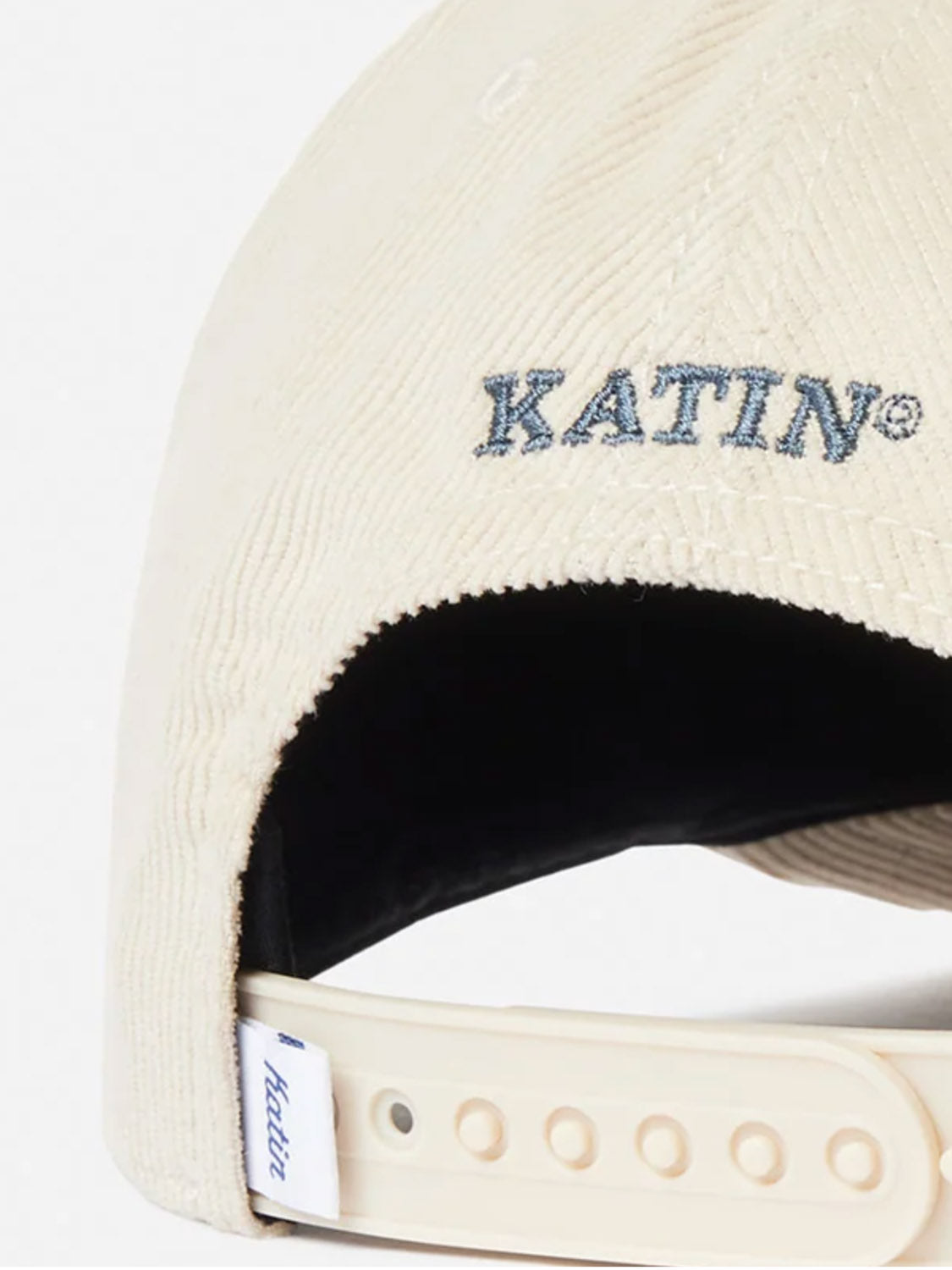 Close-up of the back of the cream-colored Katin Peaks Hat, a 5-panel cotton corduroy cap with an adjustable strap and "KATIN" embroidered in blue above the opening. A small Katin tag sits near the snapback closure.