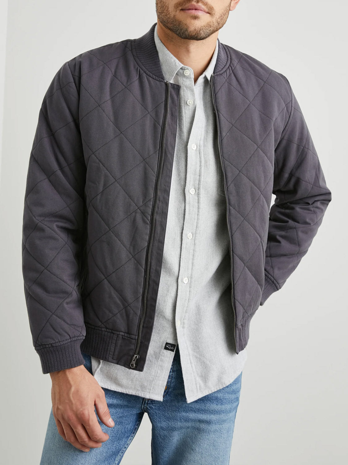 A person wearing the Rails Peninsula Jacket, a quilted bomber with ribbed collar and cuffs ideal for transitional weather, over a light gray button-up shirt and blue jeans stands with one hand in a pocket against a plain gray background.