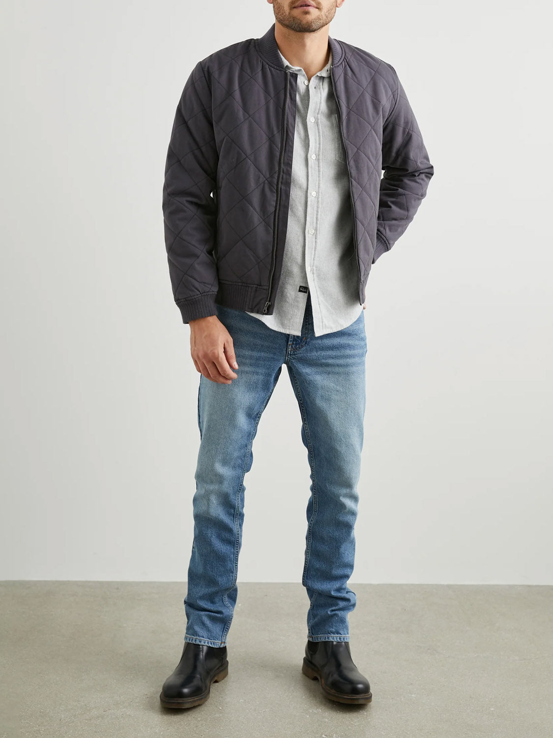 A man stands against a plain background, wearing the dark Rails Peninsula Jacket over an open gray shirt, blue jeans, and black boots. His hands rest by his sides.
