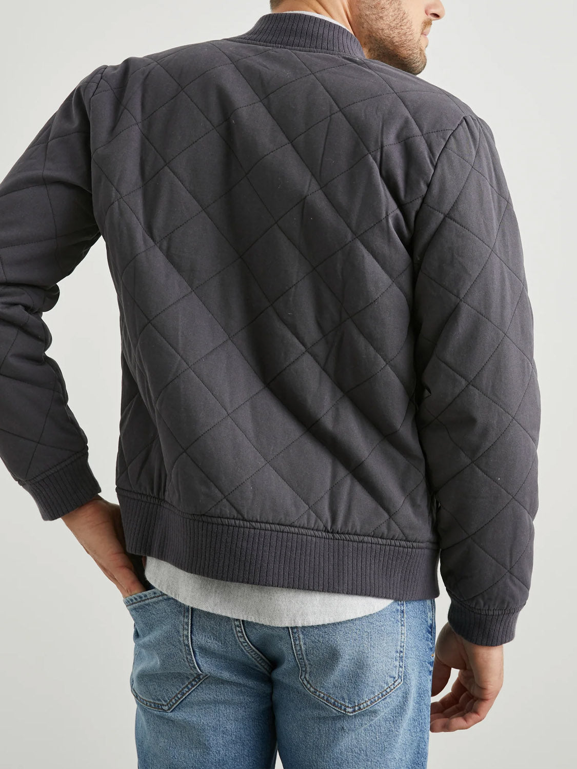 A person stands with their back to the camera, wearing the Rails Peninsula Jacket, a dark quilted bomber style with diamond-pattern stitching, paired with light blue jeans. They rest one hand on their hip against a plain light background, ready for transitional weather.
