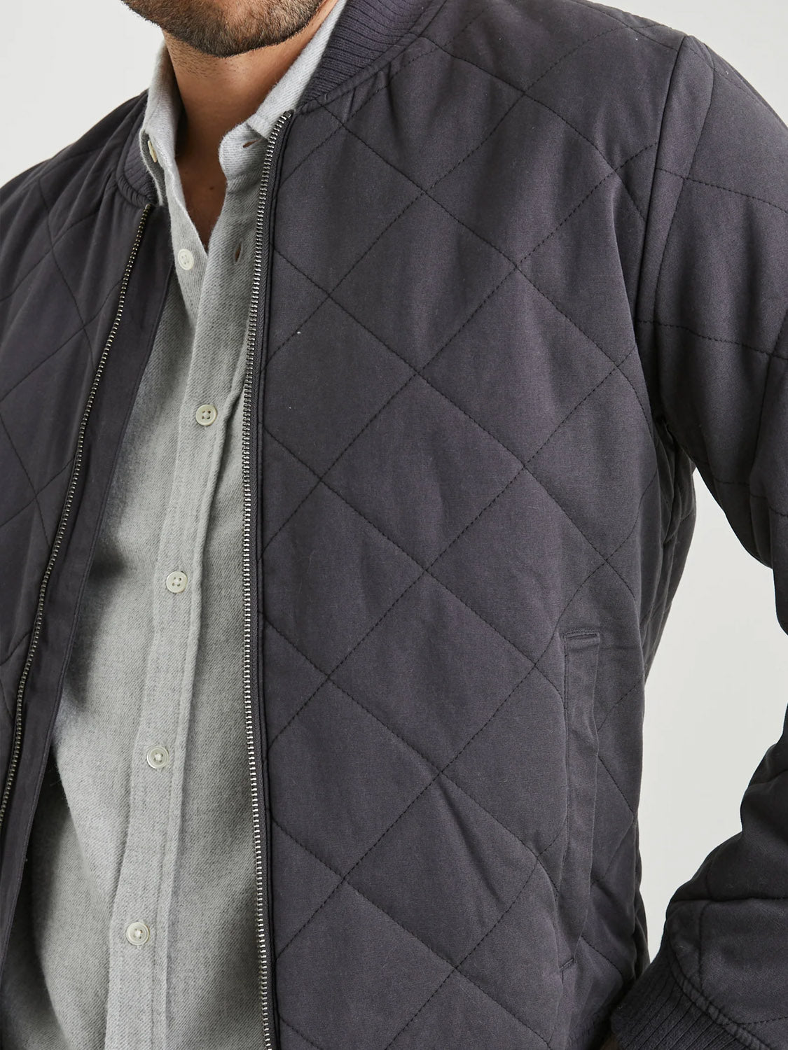 A person wears the Rails Peninsula Jacket, a stylish dark quilted bomber with diamond-pattern stitching and zipper, over a light gray button-up shirt. The head is partially visible, highlighting the jackets transitional weather appeal.
