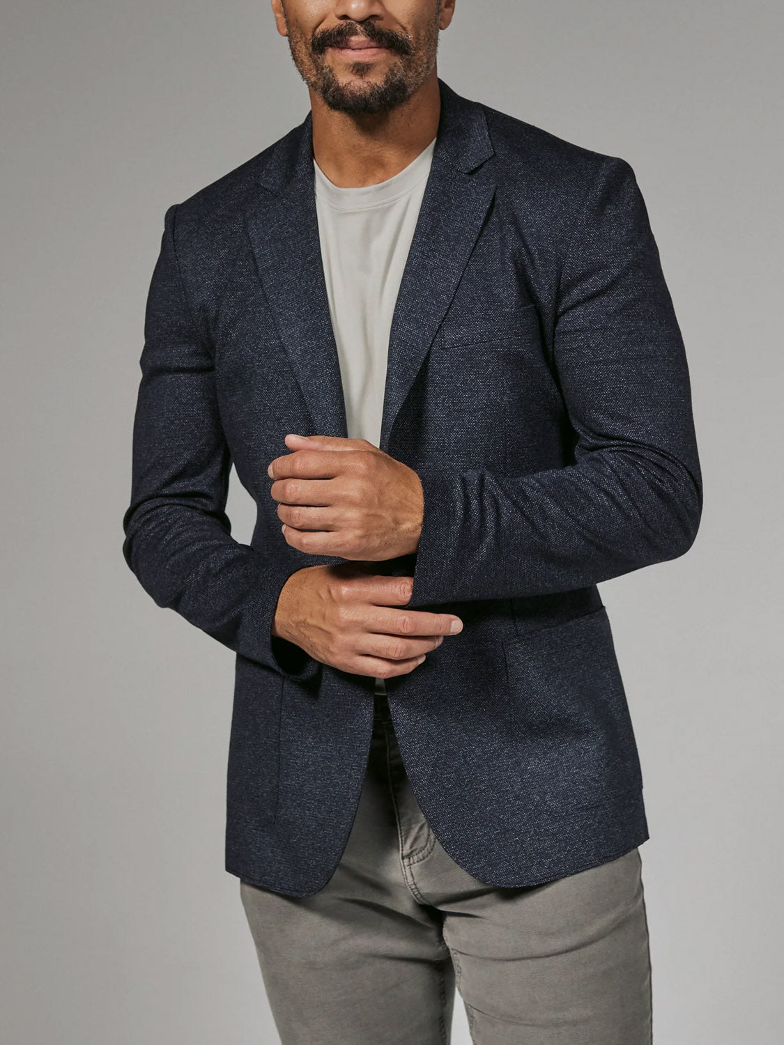 A man adjusts his sleeve while wearing the 7 Diamonds Penn Blazer by 7 Diamond over a light gray t-shirt and gray pants, standing against a plain background. His head is partially out of frame.