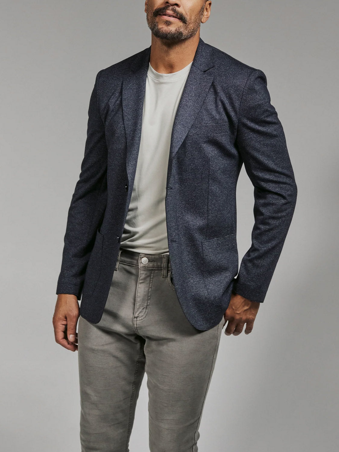 A man stands against a plain background wearing the 7 Diamonds Penn Blazer in dark blue over a light gray t-shirt and matching lightweight stretch pants, with one hand in his pocket and his face partially visible.
