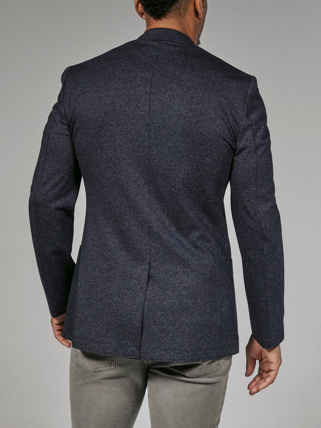 A man is seen from behind wearing the 7 Diamonds Penn Blazer by 7 Diamond, a dark navy contemporary blazer in lightweight stretch fabric, paired with light gray pants. He stands against a plain gray background.