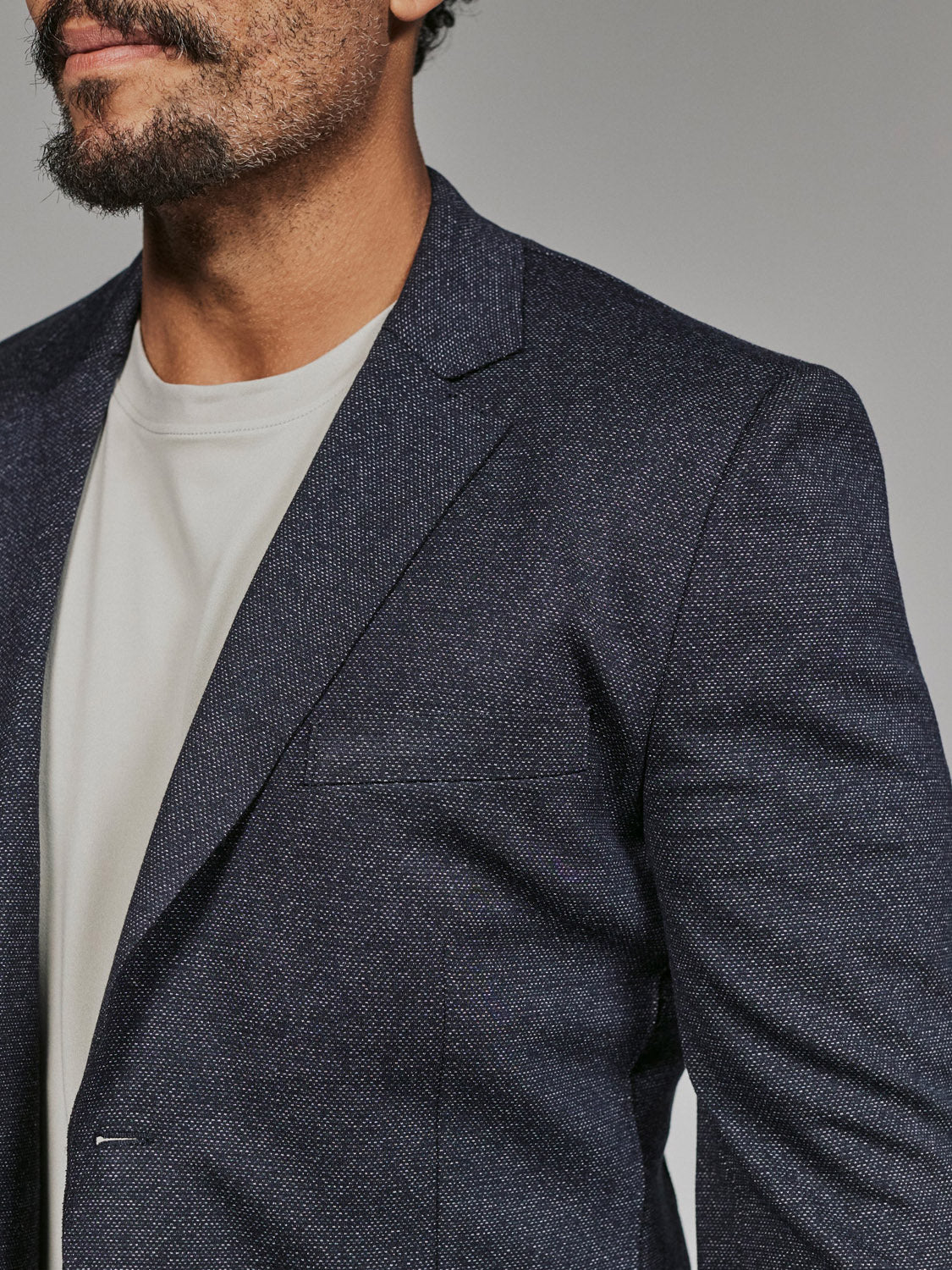 A man is shown from shoulders to mid-chest, partially revealing his face, wearing the 7 Diamonds Penn Blazer by 7 Diamond over a light crew neck shirt, with raw-edge details and a softly blurred background.