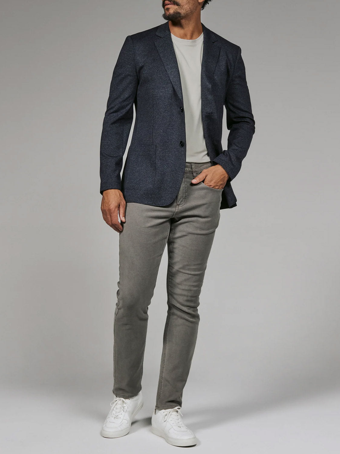 A man stands against a plain background wearing the 7 Diamonds Penn Blazer by 7 Diamond, paired with a light gray t-shirt, gray jeans, and white sneakers. His face is slightly turned and not fully visible.