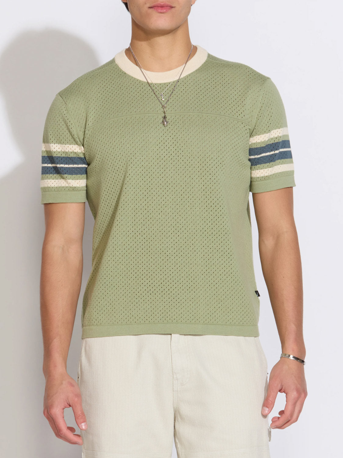 Someone stands against a plain background wearing the Civil Society Pensacola Relaxed Fit Sweater Knit Tee in light green with a cream collar and striped sleeve bands, paired with light shorts and a necklace.