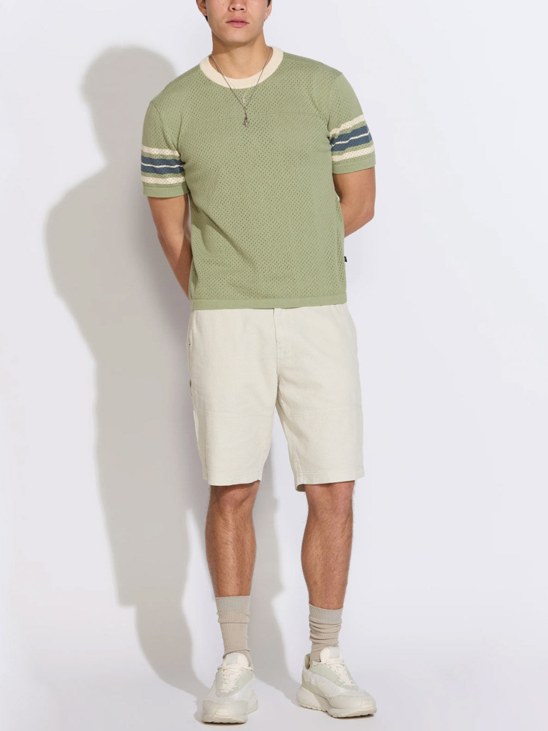 A person stands against a white background wearing the Civil Society Pensacola Relaxed Fit Sweater Knit Tee in light green, paired with beige shorts, beige socks, white sneakers, and a necklace.