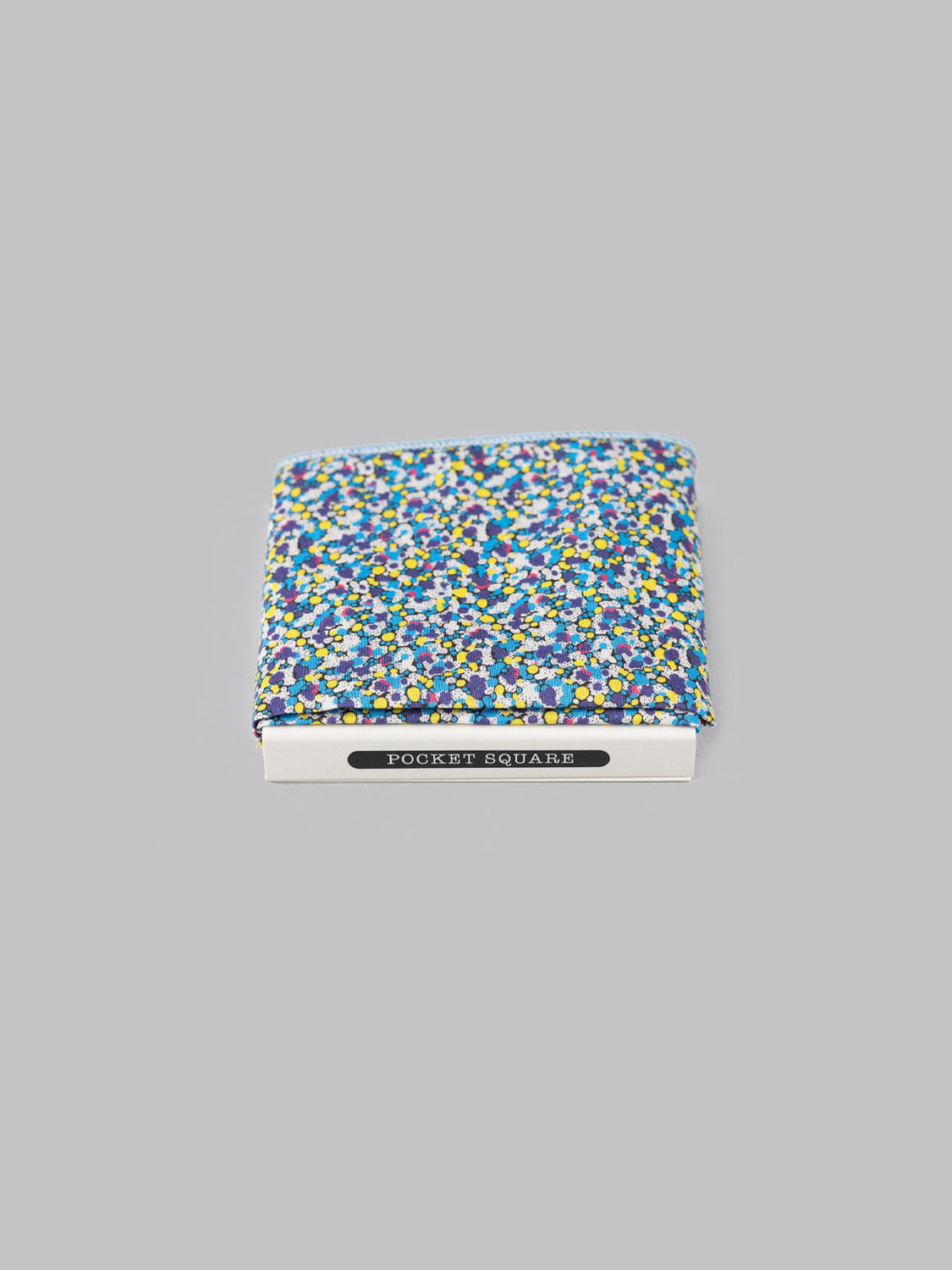 The Liberty™ Pepper Pocket Square by ID Menswear, made from Liberty of London Tana Lawn cotton in blue, yellow, and white florals, is displayed in a white POCKET SQUARE box against a plain grey background.