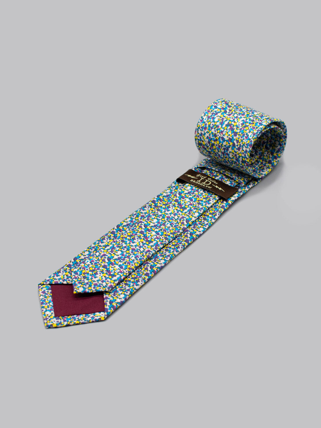 A rolled-up Liberty of London Tana Lawn™ Pepper necktie by ID Menswear, featuring a colorful floral pattern in blue, yellow, green, and white on a light gray background. The inner tip is lined with burgundy fabric.