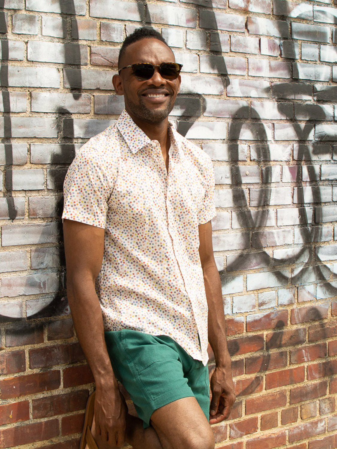 A man in sunglasses, a patterned short-sleeve shirt, and green ID Menswear Phi Phi Linen Blend Drawstring Shorts smiles while leaning against a graffiti-covered brick wall.
