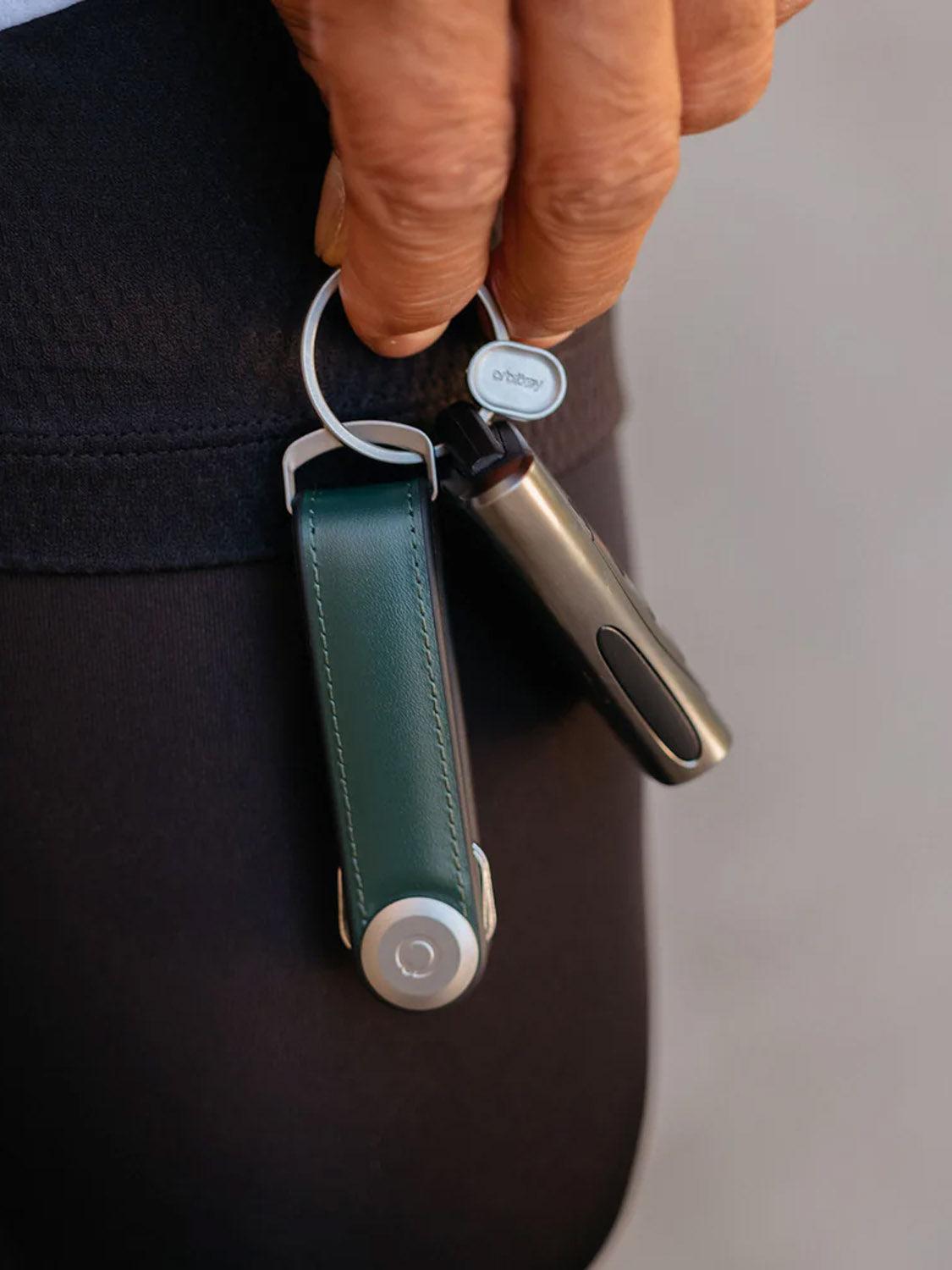 A hand holds an Orbitkeys Key Organizer Hybrid Leather in green Nappa, along with a small metallic gadget, securely clipped to dark pants.