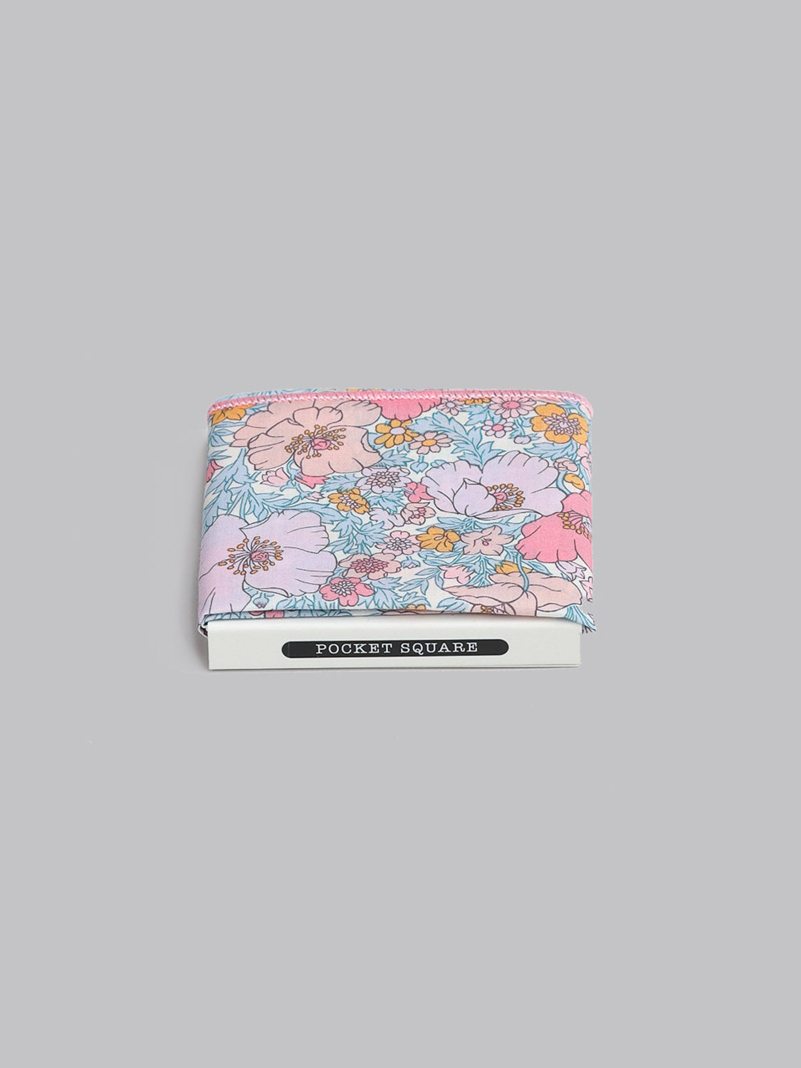 The Liberty™ Pink Lawn Pocket Square by ID Menswear, made from Tana Lawn cotton in a pastel floral pattern of pink, blue, and yellow, is shown folded on a gray background with a POCKET SQUARE label underneath.