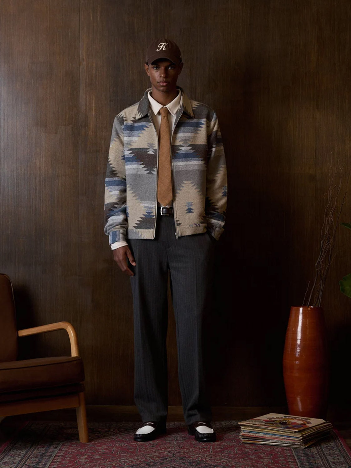 A man stands indoors by a dark wood wall, wearing a patterned jacket, white shirt, tan tie, and Kuwalla's Pinstripe Ponti Trouser with a button closure and straight leg, paired with black-and-white shoes and a brown baseball cap. A chair, vase, and magazines are nearby.