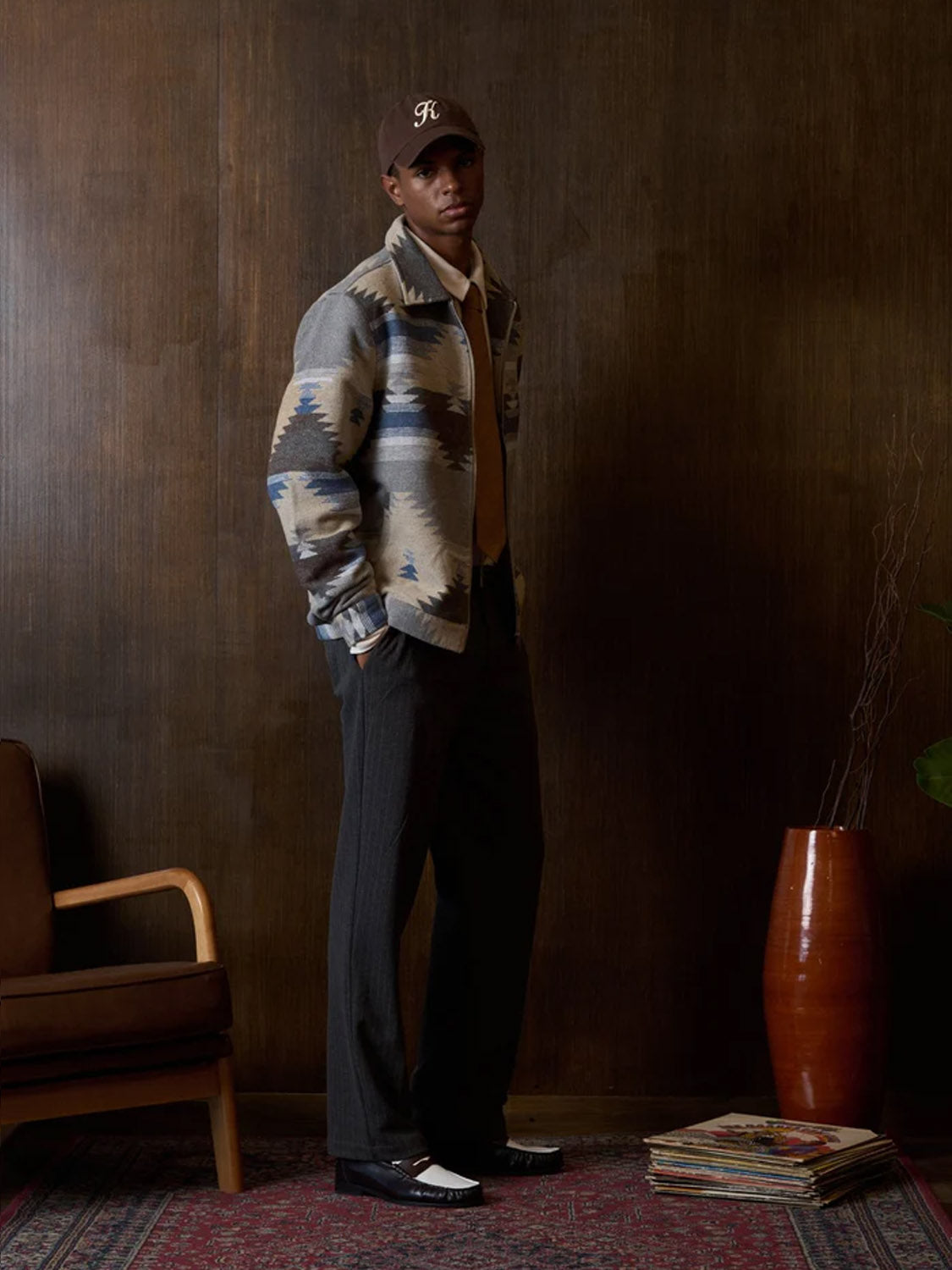A man stands indoors on a patterned rug, wearing a brown cap, patterned jacket, tan shirt, tie, black-and-white shoes, and Kuwalla Pinstripe Ponti Trousers. Beside him are a wooden chair, red vase, and a small stack of magazines.