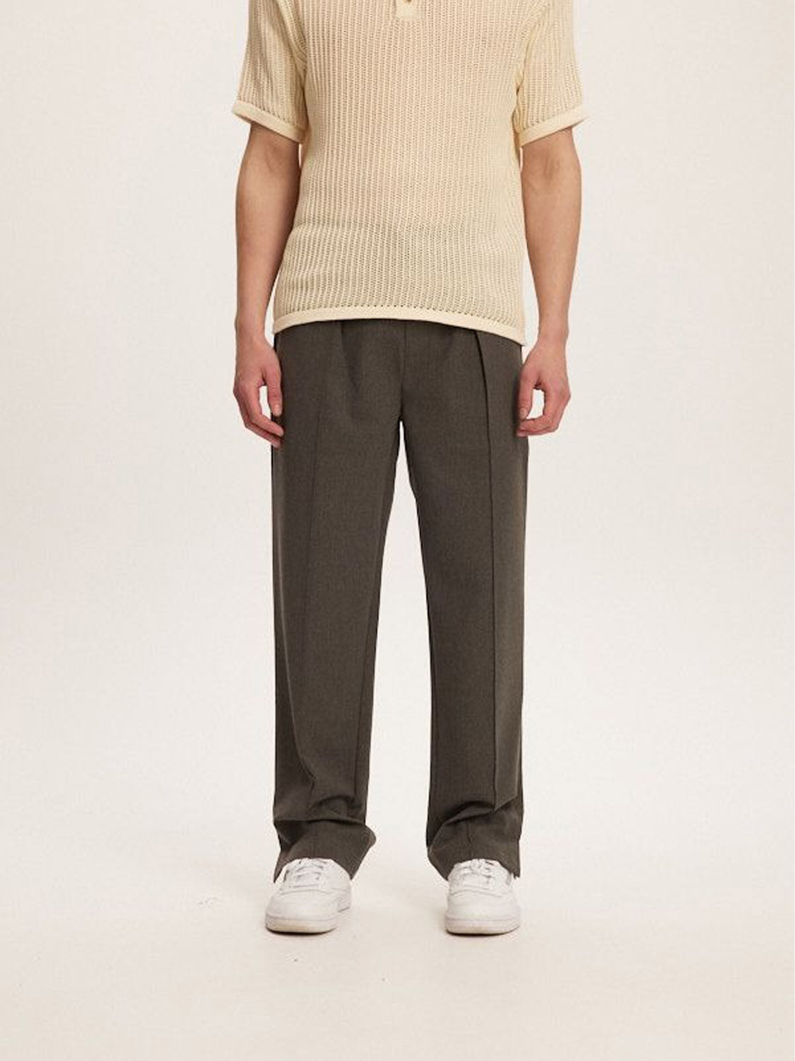 A person wearing a beige short-sleeve knit shirt, Kuwalla Pintuck Dress Pant in dark gray, and white sneakers stands against a plain background. The tailored fit of the Kuwalla ensemble offers a casual yet polished look, merging relaxed vibes with business wear charm.