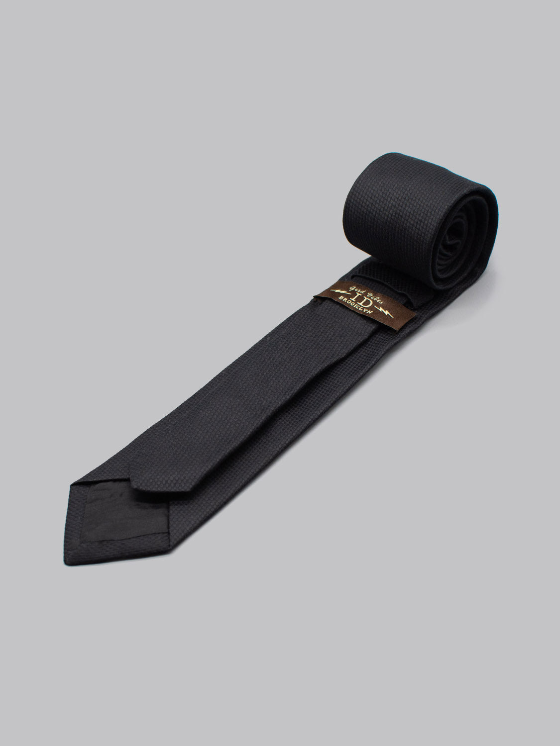 The ID Cotton Tie Pique by ID Menswear is a handmade dark cotton tie with a subtle textured pattern and small brown tag, displayed neatly rolled on a plain light gray background.