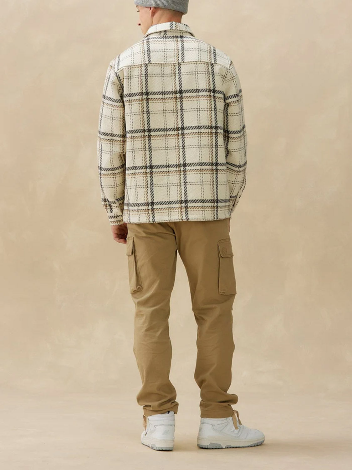 A person faces away, wearing a Kuwalla Heavyweight Plaid Shirt in beige, khaki cargo pants, white high-top sneakers, and a gray beanie against a plain beige background.