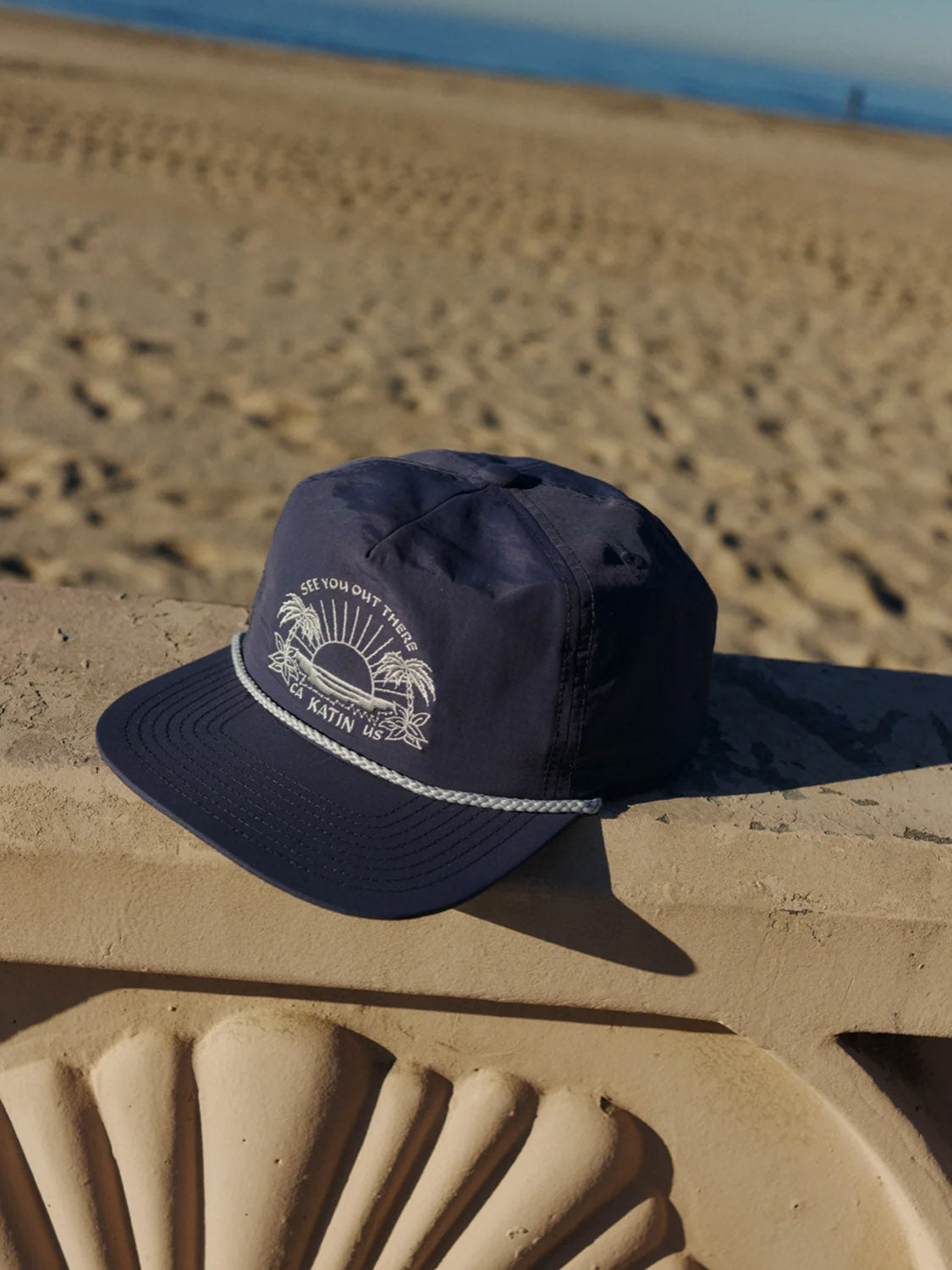 The Katin Playa Hat, a dark blue 5-panel cap featuring a white palm tree and sun design, sits on a stone ledge with a sandy beach and blue ocean in the background under a clear sky.