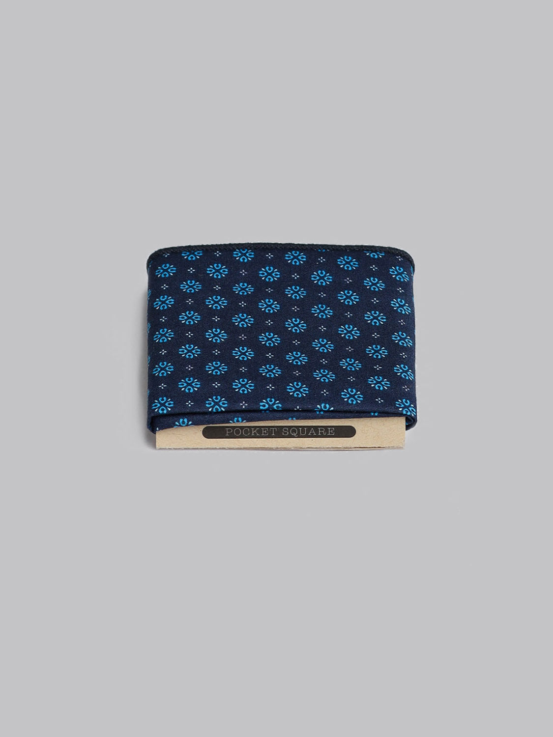 The Play Day Pocket Square by ID Menswear is handmade in Brooklyn from 100% cotton. It features a navy blue base with a small light blue floral pattern and is displayed on a branded cardboard holder against a plain grey background.