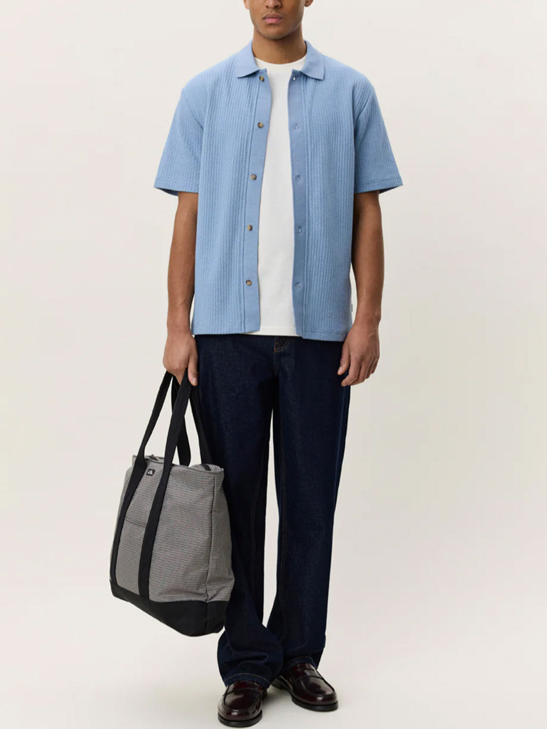 Wearing the Les Deux Pointelle SS Shirt in light blue over a white T-shirt, dark pants, and shoes, and holding a gray and black tote bag, a person stands against a plain light background.