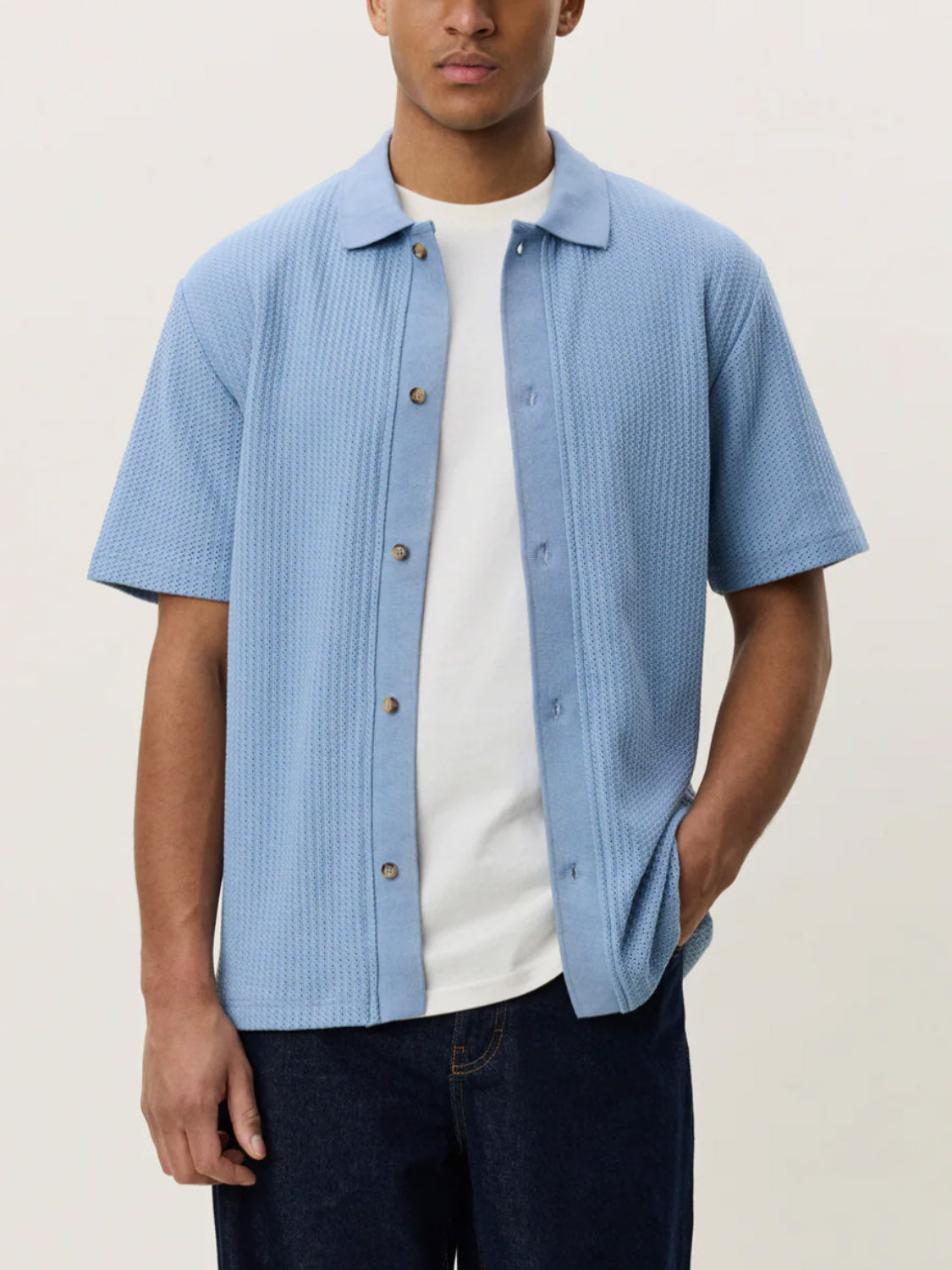 A man wearing the Les Deux Pointelle SS Shirt, a light blue camp collar button-up, over a white T-shirt and dark pants stands with one hand in his pocket against a plain background.