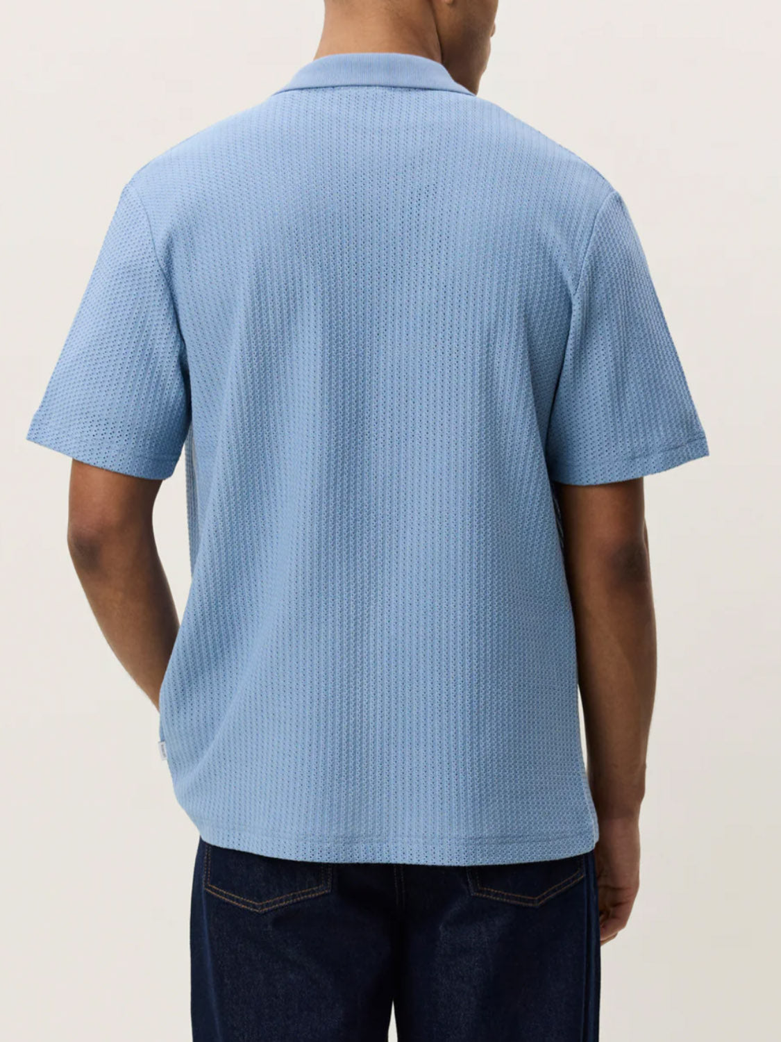 A person faces away, wearing the Les Deux Pointelle SS Shirt in light blue with short sleeves and dark blue jeans, set against a plain light background.
