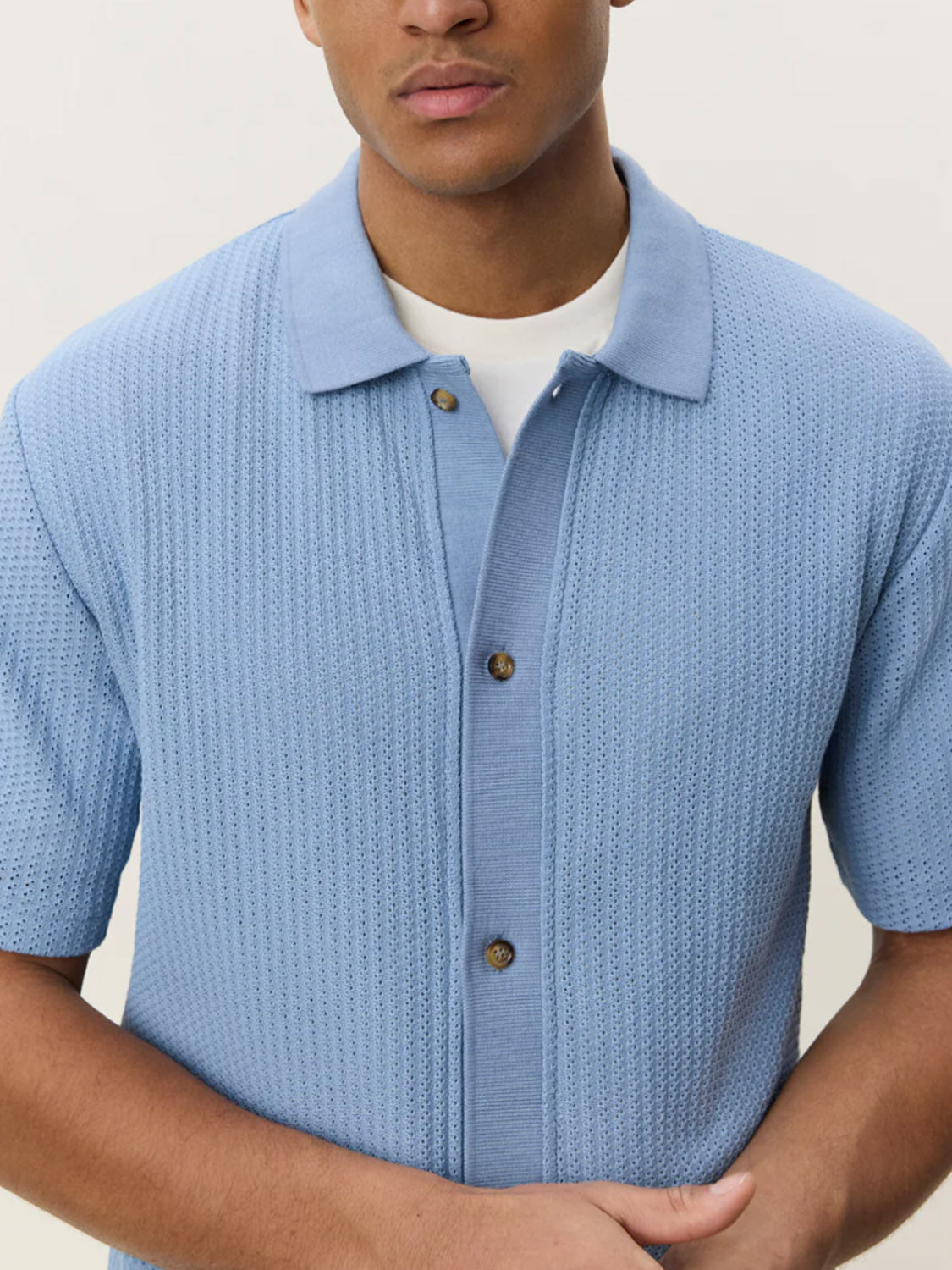 A man wears the Les Deux Pointelle SS Shirt—light blue organic cotton with a camp collar and three brown buttons—over a white tee, standing against a plain light background.