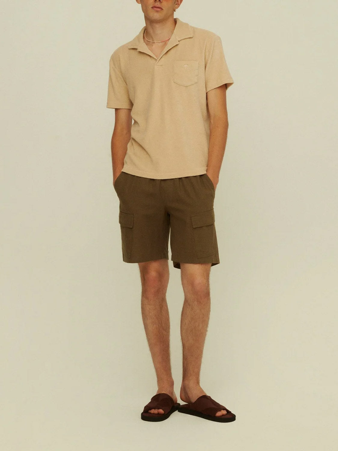 A person stands against a plain background wearing an OAS Polo Terry Shirt in beige cotton terrycloth, paired with brown cargo shorts and dark brown sandals, hands in their pockets for a comfortable, breathable look.