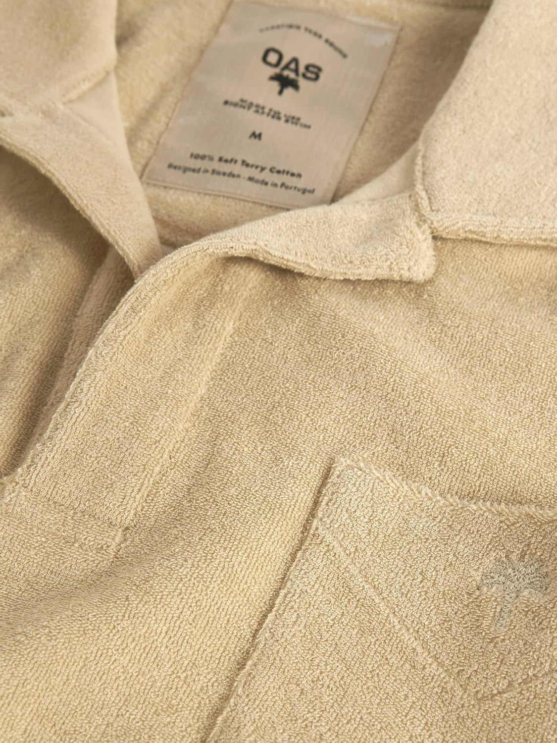 Close-up of the OAS Polo Terry Shirt in beige cotton terrycloth, featuring a chest pocket, collar, and OAS label with size details. The soft, textured fabric is breathable for all-day comfort.