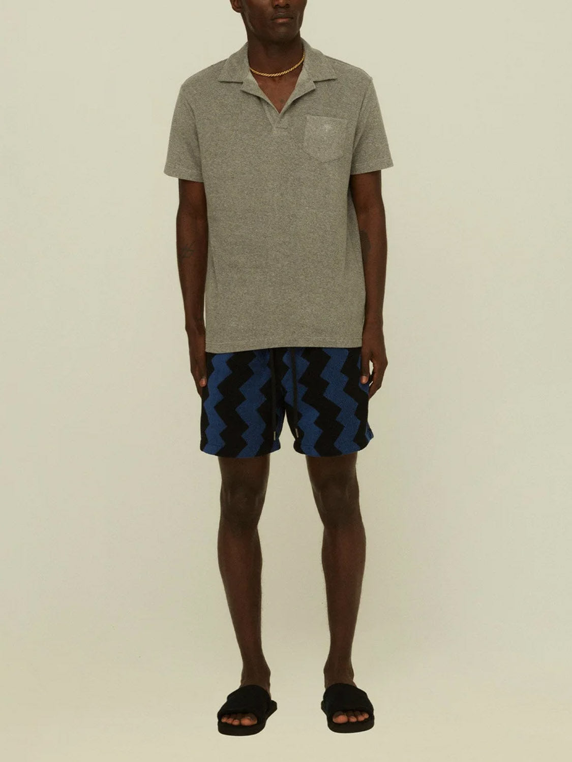 A person stands against a plain background wearing an OAS Polo Terry Shirt, featuring short sleeves and breathable cotton terrycloth, paired with blue and black zigzag shorts, black slide sandals, and a gold necklace.