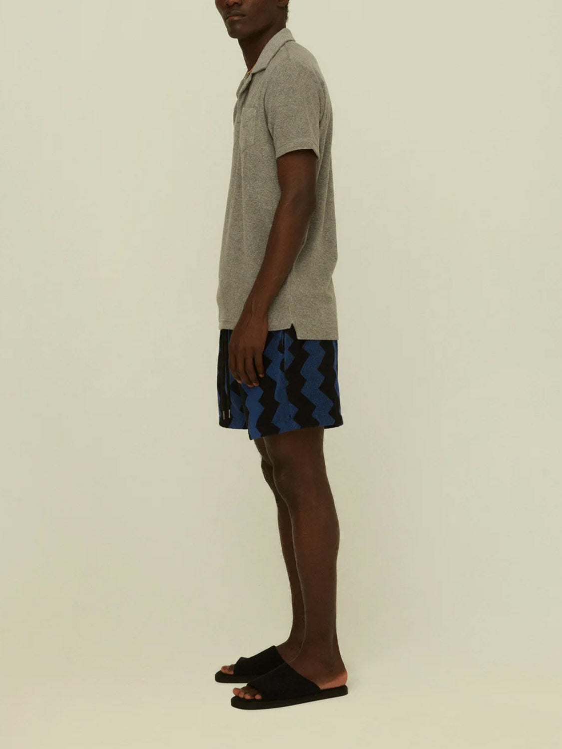 A man stands sideways against a plain light background wearing the OAS Polo Terry Shirt in breathable gray cotton terrycloth, blue and black zigzag shorts, and black slide sandals.
