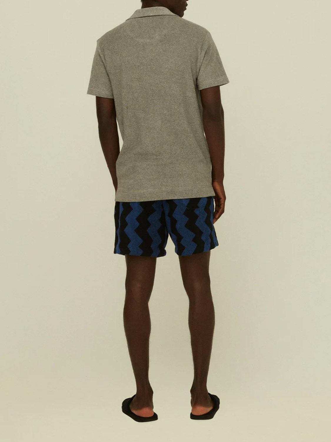 A person faces away, wearing the OAS Polo Terry Shirt in breathable grey cotton terrycloth, paired with zigzag patterned shorts and black flip-flops against a plain light background.