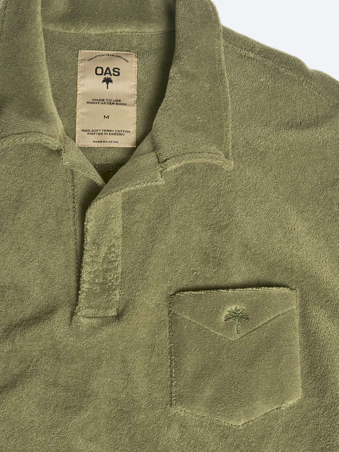 The OAS Polo Terry Shirt in olive green cotton terrycloth features a collar, partial button placket, chest pocket with embroidered palm tree, breathable fabric, and an inside label with size and garment details.