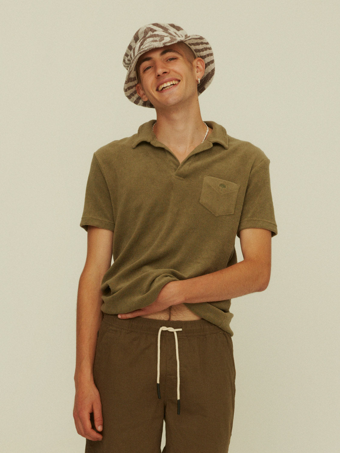 A young man smiles with one hand in his OAS Polo Terry Shirt in olive green, paired with brown drawstring pants and a patterned bucket hat, set against a plain beige background.