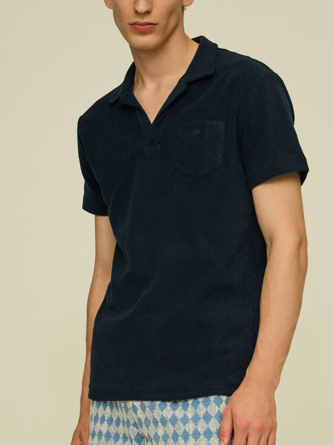 A man stands against a plain light background wearing an OAS Polo Terry Shirt with a chest pocket and light blue patterned shorts. The breathable cotton terrycloth offers comfort, and his head is partially out of frame.