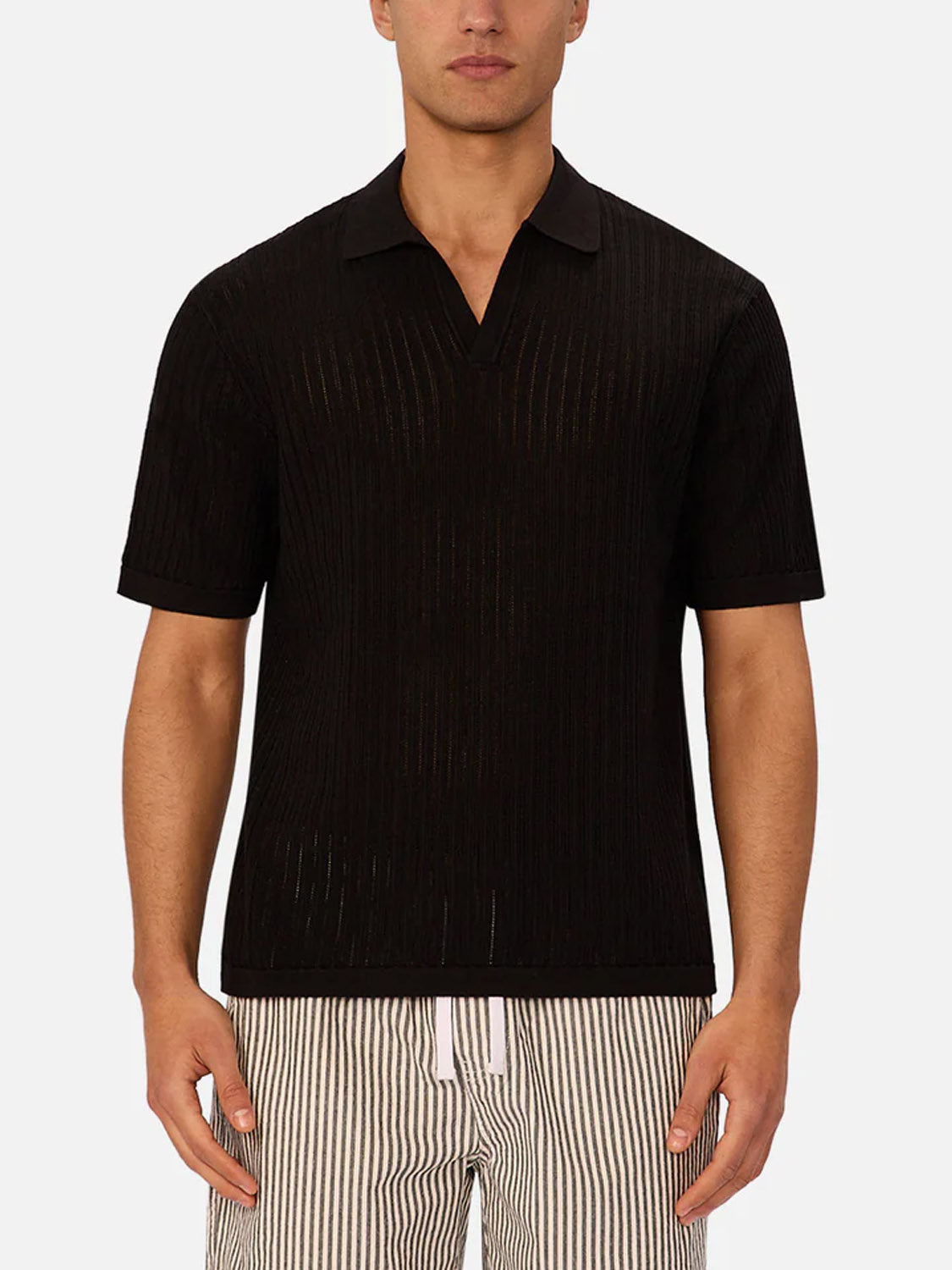 A man wears the Industrie Poolside Polo in black, made from breathable cotton with a ribbed knit and relaxed collar, paired with white drawstring, vertically striped shorts, standing against a plain light grey background.